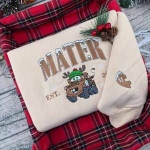 Puede incluir: Una sudadera beige con la palabra "MATER" bordada en marr&oacute;n, con un dise&ntilde;o de personaje de dibujos animados. La sudadera est&aacute; doblada dentro de una cesta a cuadros roja y negra, con adornos de pino y bayas.