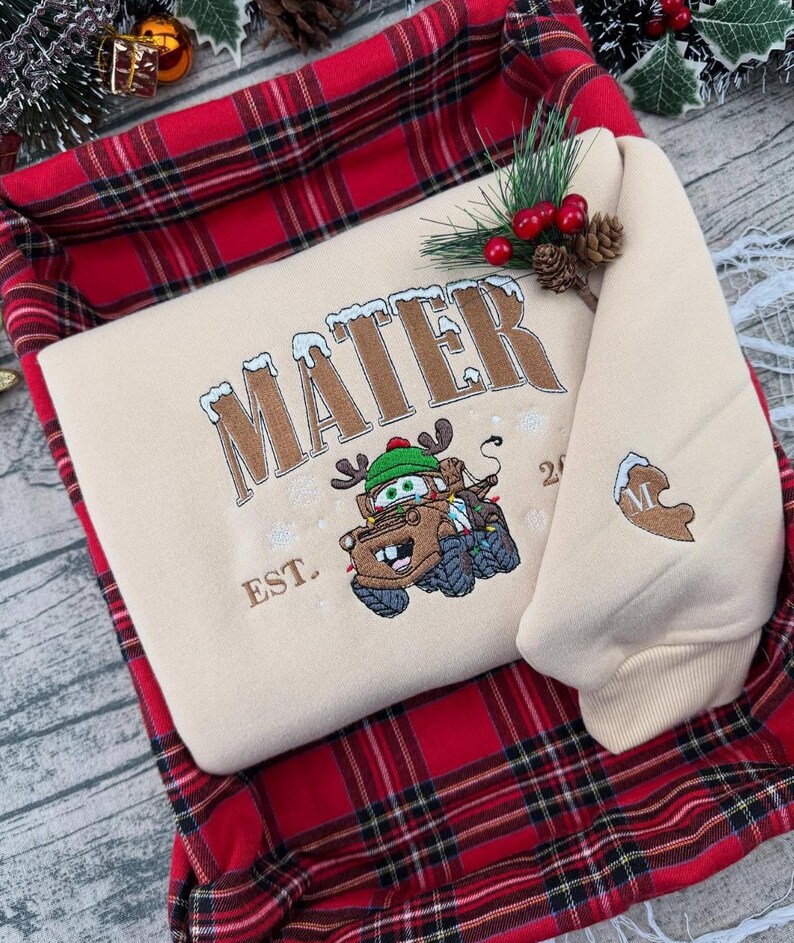 Puede incluir: Una sudadera beige con la palabra "MATER" bordada en marr&oacute;n, con un dise&ntilde;o de personaje de dibujos animados. La sudadera est&aacute; doblada dentro de una cesta a cuadros roja y negra, con adornos de pino y bayas.