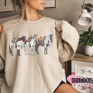 May include: A beige sweatshirt with a graphic print of four horses with saddles. The horses are brown, white, and black.
