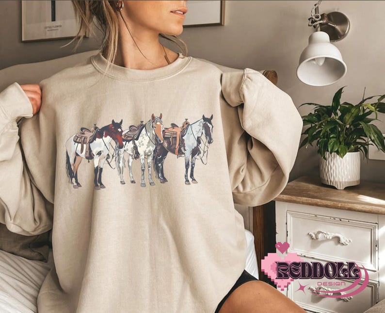 May include: A beige sweatshirt with a graphic print of four horses with saddles. The horses are brown, white, and black.