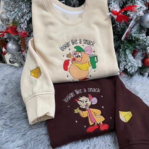 Könnte beinhalten: Zwei Sweatshirts, eines cremefarben und eines braun, mit gestickten Cartoon-Mäusen. Das cremefarbene Sweatshirt hat eine Maus, die ein Getränk hält, und den Text "lookin' like a snack". Das braune Sweatshirt hat eine Maus, die Käse hält. Beide haben eine Käse-Grafik.