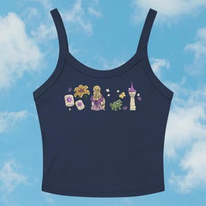 May include: Navy blue tank top with a colorful graphic featuring a blonde character in a purple dress, a green chameleon, a tower, and floating lanterns. The design includes floral and butterfly accents, set against a blue sky background.