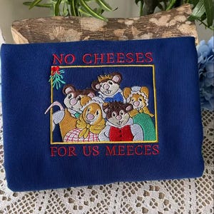 May include: Navy blue sweatshirt with an embroidered design featuring a group of mice inside a gold frame. The text "NO CHEESES FOR US MEECE" is embroidered in red. The design includes green and red accents.