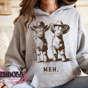 K&ouml;nnte beinhalten: Grauer Hoodie mit einem Aufdruck von zwei Ziegen, die Cowboyh&uuml;te tragen. Die Ziegen sind braun und wei&szlig; und der Text "MEH." ist darunter gedruckt.