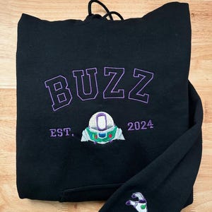 May include: Black hoodie with the word "BUZZ" embroidered in purple, along with a Buzz Lightyear design and the year "2024." The sleeve features a matching embroidered design. The hoodie has a drawstring hood.