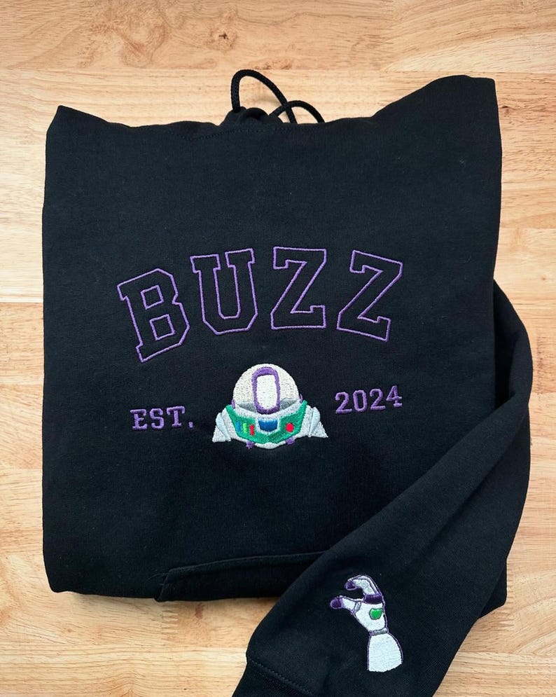 May include: Black hoodie with the word "BUZZ" embroidered in purple, along with a Buzz Lightyear design and the year "2024." The sleeve features a matching embroidered design. The hoodie has a drawstring hood.