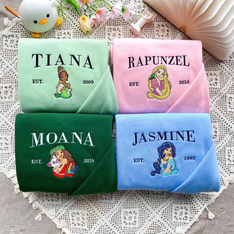 Może przedstawiać: Cztery złożone bluzy w pastelowych kolorach: jasna zieleń, r&oacute;ż, ciemna zieleń i jasny błękit. Każda z nich ma wyszyte czarnym kolorem imię księżniczki, wraz z ilustracją i rokiem założenia. Księżniczki to Tiana, Roszpunka, Moana i Jasmine.