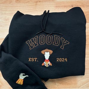 May include: Black hoodie with the name "WOODY" embroidered in brown, along with an image of Woody and the text "EST. 2024." The left sleeve features an embroidered image of Woody's hand. The hoodie has a drawstring hood.