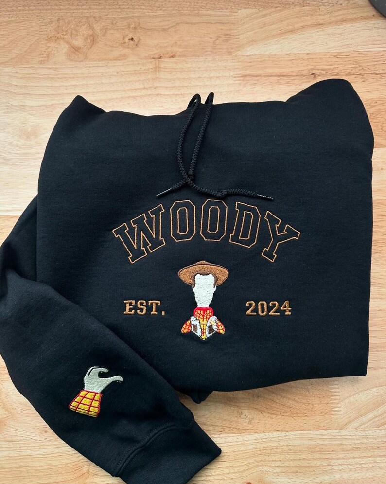 May include: Black hoodie with the name "WOODY" embroidered in brown, along with an image of Woody and the text "EST. 2024." The left sleeve features an embroidered image of Woody's hand. The hoodie has a drawstring hood.
