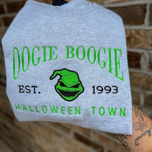 K&ouml;nnte beinhalten: Hellgraues Sweatshirt mit gr&uuml;n gesticktem Text und Grafik. Der Text lautet "Oogie Boogie", "EST. 1993" und "Halloween Town". Die Grafik zeigt ein gr&uuml;nes Gesicht mit einem finsteren L&auml;cheln.