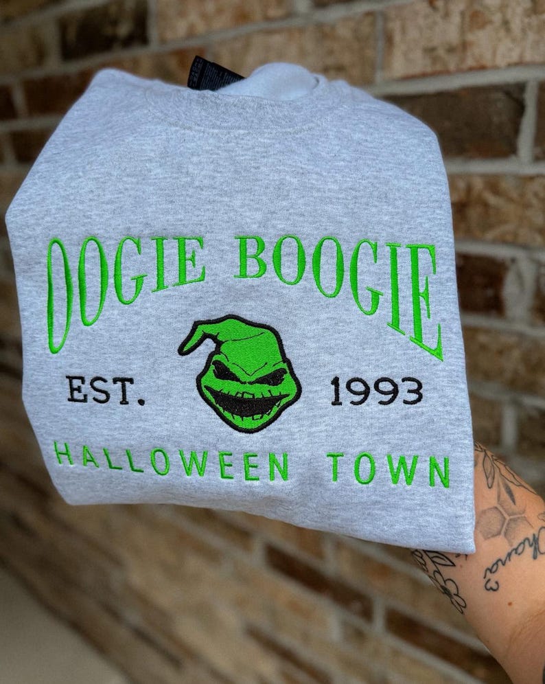 K&ouml;nnte beinhalten: Hellgraues Sweatshirt mit gr&uuml;n gesticktem Text und Grafik. Der Text lautet "Oogie Boogie", "EST. 1993" und "Halloween Town". Die Grafik zeigt ein gr&uuml;nes Gesicht mit einem finsteren L&auml;cheln.