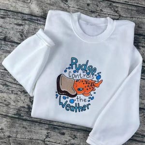 K&ouml;nnte beinhalten: Wei&szlig;es Sweatshirt mit Rundhalsausschnitt und langen &Auml;rmeln. Auf der Vorderseite befindet sich eine farbenfrohe Grafik eines orangefarbenen Fisches mit dem Text "Pudge controls the weather" in Blau und Schwarz.