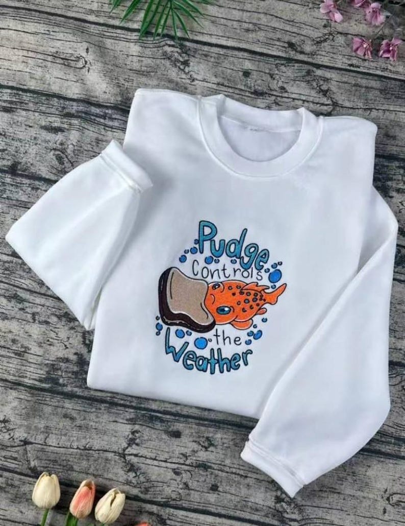 K&ouml;nnte beinhalten: Wei&szlig;es Sweatshirt mit Rundhalsausschnitt und langen &Auml;rmeln. Auf der Vorderseite befindet sich eine farbenfrohe Grafik eines orangefarbenen Fisches mit dem Text "Pudge controls the weather" in Blau und Schwarz.
