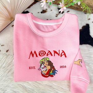 May include: A light pink crewneck sweatshirt featuring embroidered red text that reads MOANA, with the text EST. 2016 on either side. Below the text is an embroidered illustration of a character holding a pig and a rooster. A hand heart is on the sleeve.