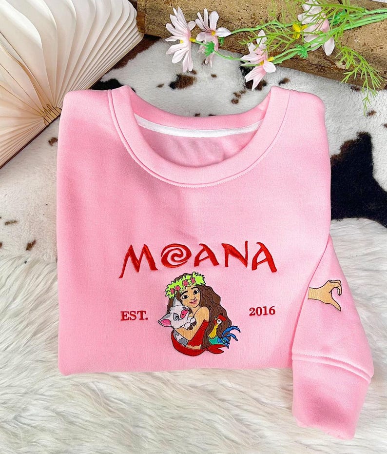 May include: A light pink crewneck sweatshirt featuring embroidered red text that reads MOANA, with the text EST. 2016 on either side. Below the text is an embroidered illustration of a character holding a pig and a rooster. A hand heart is on the sleeve.