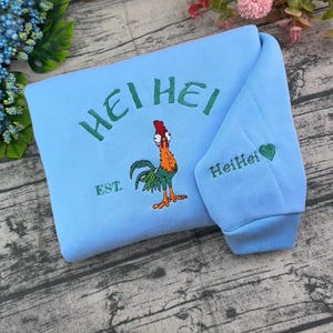Pode incluir: Uma camisola azul-clara com um gr&aacute;fico bordado de um galo colorido com o texto HEI HEI e EST. acima e abaixo da personagem. A manga inclui o texto bordado HeiHei com um pequeno s&iacute;mbolo de cora&ccedil;&atilde;o verde.