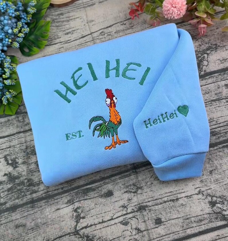 Pode incluir: Uma camisola azul-clara com um gr&aacute;fico bordado de um galo colorido com o texto HEI HEI e EST. acima e abaixo da personagem. A manga inclui o texto bordado HeiHei com um pequeno s&iacute;mbolo de cora&ccedil;&atilde;o verde.