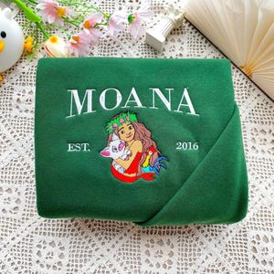 Może przedstawiać: Ciemnozielona bluza z haftowanym białym napisem "MOANA". Poniżej znajduje się haftowany obraz kobiety trzymającej świnię i koguta. Na bluzie wyhaftowano r&oacute;wnież napis "EST. 2016".