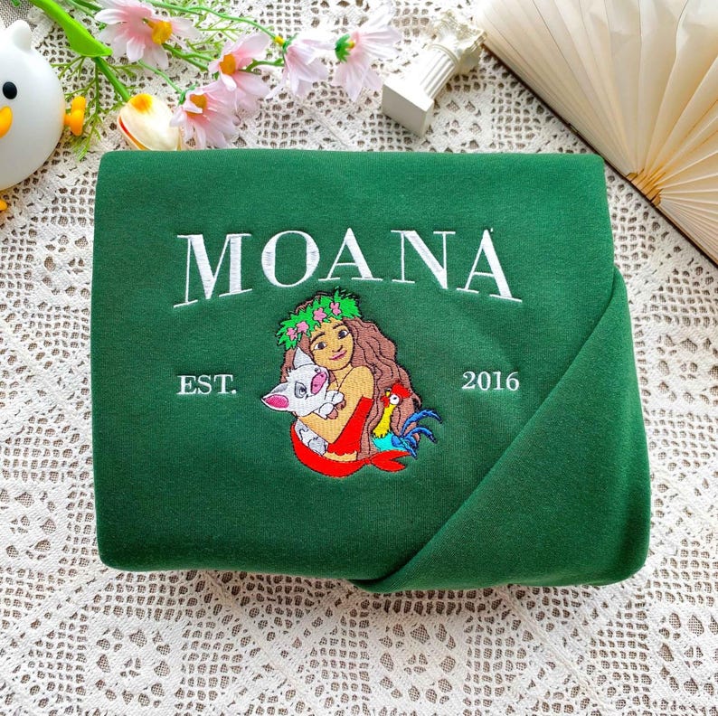Może przedstawiać: Ciemnozielona bluza z haftowanym białym napisem "MOANA". Poniżej znajduje się haftowany obraz kobiety trzymającej świnię i koguta. Na bluzie wyhaftowano r&oacute;wnież napis "EST. 2016".