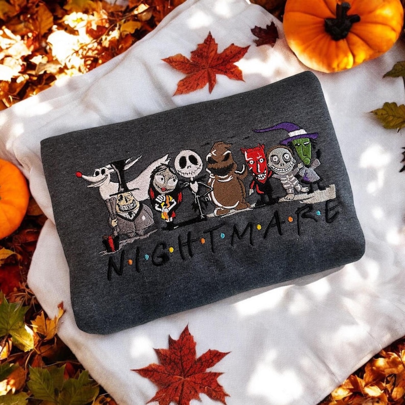 May include: Dark gray sweatshirt featuring an embroidered design of characters from the movie "The Nightmare Before Christmas." The word "NIGHTMARE" is embroidered below the characters in black. The sweatshirt is folded on a white surface with autumn leaves and pumpkins.