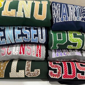 Può includere: Una pila di otto felpe piegate in vari colori, tra cui verde, blu navy e grigio. Ogni felpa presenta grandi lettere in tessuto applicate con motivi diversi come vichy, fiori e pois, che mostrano il testo PLNU, MARIETTA, GENESEO, PSU, WISCONSIN, TARLETON, WLL e SDSU.