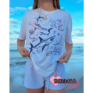 K&ouml;nnte beinhalten: Wei&szlig;es T-Shirt mit einem grafischen Druck verschiedener Meeresbewohner, darunter Haie, Quallen und ein Tintenfisch. Das Shirt wird von einer Person getragen, die am Strand steht.