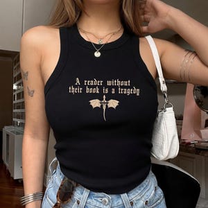 Peut inclure: Débardeur noir côtelé avec l'inscription "A reader without their book is a tragedy" et un motif de dragon. Le haut sans manches a un col rond. Le texte et le motif sont de couleur beige clair.