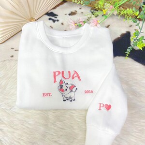 May include: A white crewneck sweatshirt featuring an embroidered graphic of a small pig character. The text PUA is stitched in pink above the pig, with EST. on the left and 2016 on the right. The sleeve features the letter P and a pink heart.