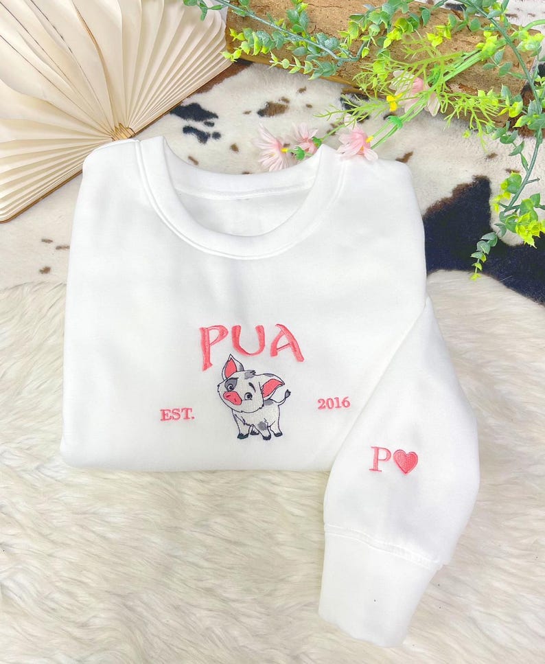 May include: A white crewneck sweatshirt featuring an embroidered graphic of a small pig character. The text PUA is stitched in pink above the pig, with EST. on the left and 2016 on the right. The sleeve features the letter P and a pink heart.