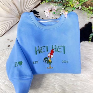 May include: A light blue crewneck sweatshirt featuring green embroidered text that reads HEI HEI, with an embroidered graphic of a rooster with a red comb and yellow body, the text EST. 2016, and an H with a green heart on the left sleeve.