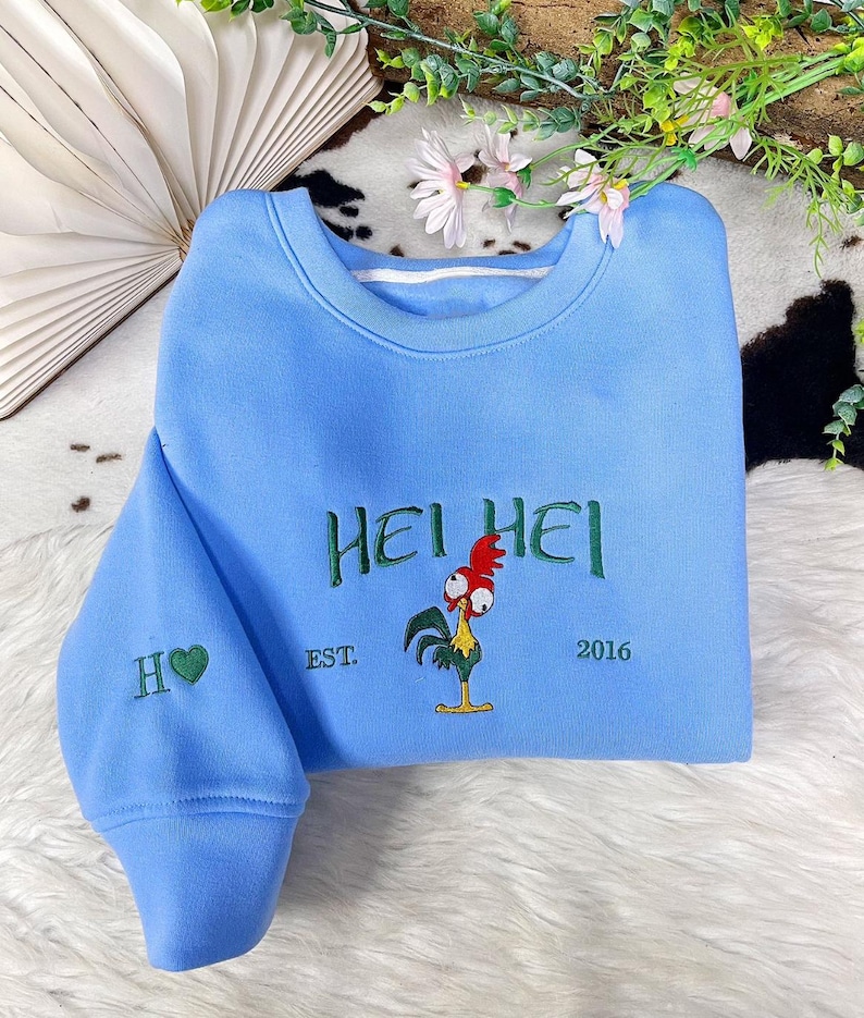 May include: A light blue crewneck sweatshirt featuring green embroidered text that reads HEI HEI, with an embroidered graphic of a rooster with a red comb and yellow body, the text EST. 2016, and an H with a green heart on the left sleeve.