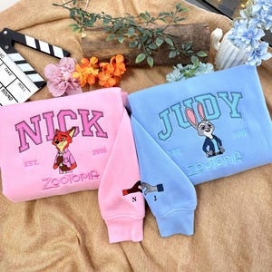 May include: Two sweatshirts, one pink and one light blue, featuring embroidered designs. The pink sweatshirt says "NICK" with a fox illustration, and the blue one says "JUDY" with a bunny. Both have "Zootopia" and "2016" on them.