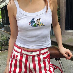 May include: White vest top featuring a cartoon image of Stitch and Lilo. The top is worn with red and white striped shorts. A casual outfit, perfect for warmer weather.