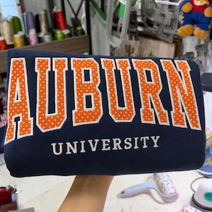Könnte beinhalten: Ein marineblaues Sweatshirt mit dem Wort AUBURN in großen, orangefarbenen Blockbuchstaben mit weißen Punkten und weißen Umrandungen sowie dem Wort UNIVERSITY in kleineren, weißen, serifenlosen Großbuchstaben direkt darunter.