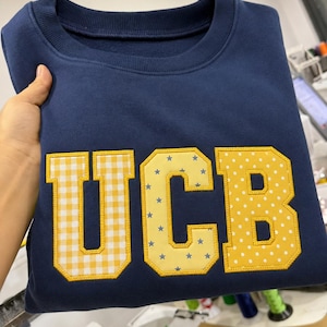 Può includere: Felpa blu navy con le lettere "UCB" applicate sul davanti. La "U" è a quadretti gialli, la "C" è gialla con stelle blu e la "B" è gialla con pois bianchi. Ha uno scollo tondo.