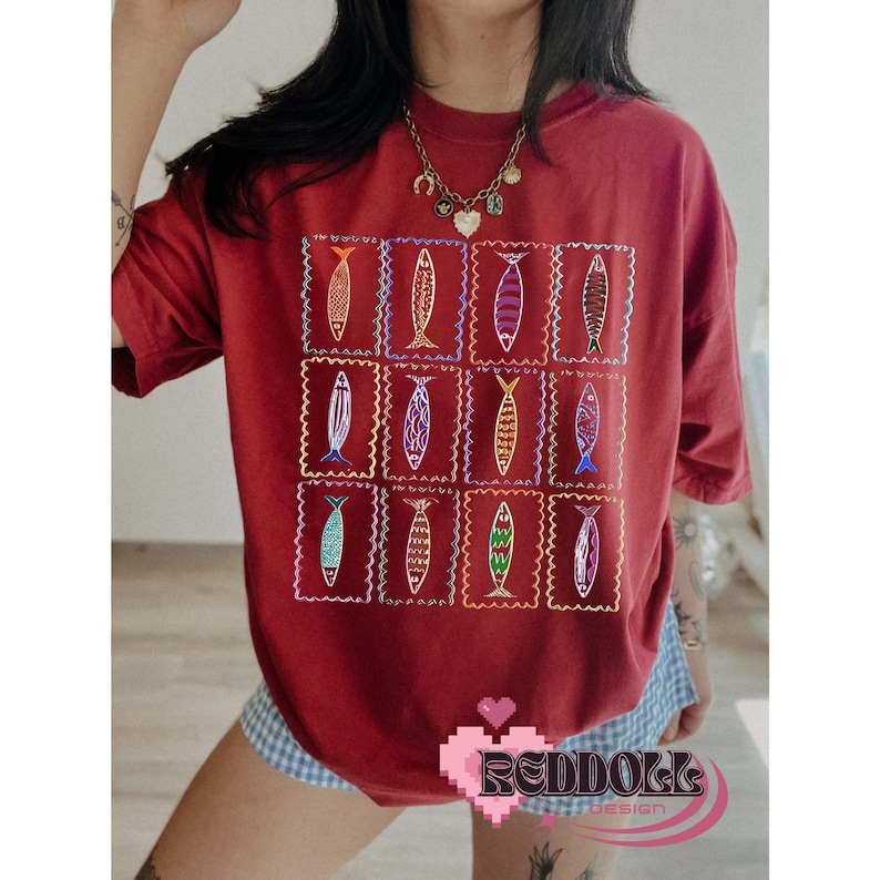 May include: A red t-shirt with a pattern of colorful fish in squares. The fish are embroidered on the shirt. The shirt is being worn by a person.