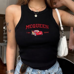 May include: Black ribbed tank top with the embroidered text "MCQUEEN" in red above a cartoon car graphic. The text "i am speed" is below the car. The top is sleeveless and has a scoop neckline.
