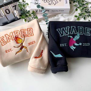 May include: Two sweatshirts, one beige and one black, with embroidered designs. The beige sweatshirt features the name "EMBER" in orange, a cartoon character, and "EST. 2023." The black sweatshirt displays "WADE" in teal, a cartoon character, and "EST. 2023."