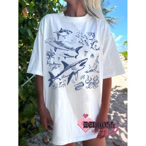 K&ouml;nnte beinhalten: Wei&szlig;es T-Shirt mit einem blauen Grafikdruck von verschiedenen Haien, einem Oktopus und anderen Meeresbewohnern. Die Grafik ist mittig auf dem Shirt platziert und bedeckt den gr&ouml;&szlig;ten Teil der Vorderseite.