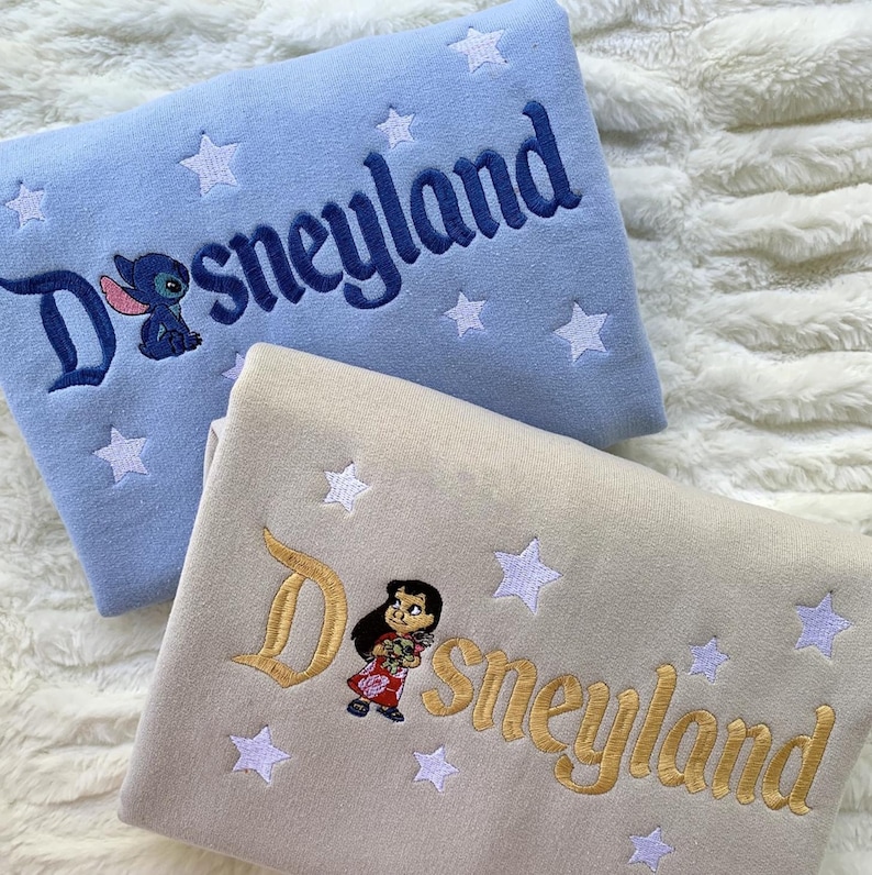 May include: Two sweatshirts, one light blue and one beige, embroidered with "Disneyland" in script. The light blue sweatshirt features Stitch, and the beige one features Lilo. Both have white star accents.