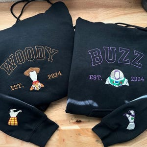 May include: Two black hoodies with embroidered designs. One features "WOODY" in gold lettering with a Woody character and "EST. 2024". The other says "BUZZ" in purple with a Buzz Lightyear design and "EST. 2024". Sleeves have matching character details.