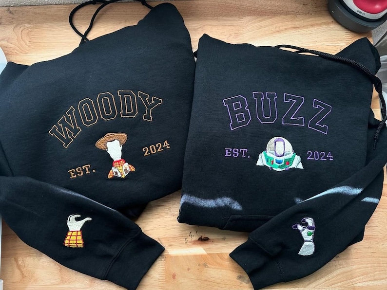 May include: Two black hoodies with embroidered designs. One features "WOODY" in gold lettering with a Woody character and "EST. 2024". The other says "BUZZ" in purple with a Buzz Lightyear design and "EST. 2024". Sleeves have matching character details.