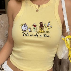 Puede incluir: Una camiseta de tirantes de color amarillo pálido con un bordado que muestra una tetera, una taza, una rosa roja en una cúpula de cristal, una figura femenina con vestido amarillo y un candelabro. El texto "Tale as old as time" está bordado en negro.