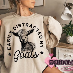 May include: A beige sweatshirt with the text "Easily Distracted By Goats" in black lettering. A black and white illustration of a goat's head is centered on the sweatshirt.