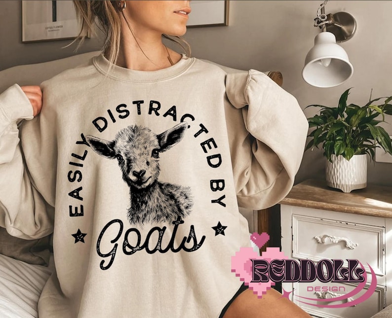 May include: A beige sweatshirt with the text "Easily Distracted By Goats" in black lettering. A black and white illustration of a goat's head is centered on the sweatshirt.