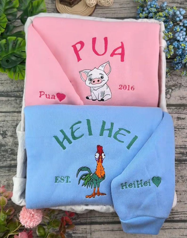 Pode incluir: Uma camisola rosa com um porquinho bordado, o texto PUA e 2016, ao lado de uma camisola azul clara com um galo bordado, o texto HEI HEI e EST. Ambas as pe&ccedil;as incluem pequenos bordados de cora&ccedil;&otilde;es e nomes nas mangas.