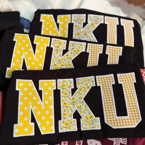 May include: Three black t-shirts featuring the letters NKU in large varsity-style applique. Each letter has a white border. The N is yellow with white polka dots, the K is a yellow floral print, and the U is a yellow and white gingham check.