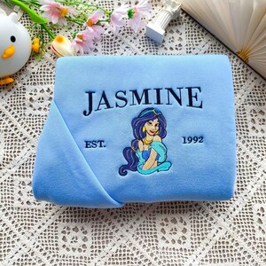 Może przedstawiać: Jasnoniebieski koc z haftowanym granatowym napisem "JASMINE". Poniżej znajduje się haftowany wizerunek kobiety z kresk&oacute;wki z ciemnymi włosami i złotą biżuterią. Na kocu widnieje r&oacute;wnież napis "EST. 1992".