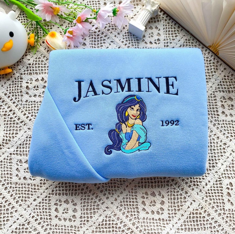 Może przedstawiać: Jasnoniebieski koc z haftowanym granatowym napisem "JASMINE". Poniżej znajduje się haftowany wizerunek kobiety z kresk&oacute;wki z ciemnymi włosami i złotą biżuterią. Na kocu widnieje r&oacute;wnież napis "EST. 1992".