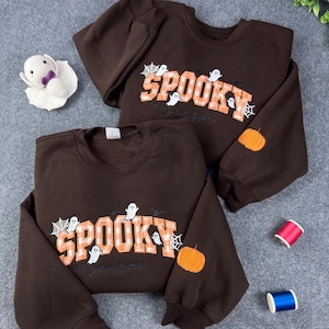 Peut inclure: Deux sweat-shirts à col rond marron foncé avec le mot SPOOKY en lettres à carreaux orange et blanc. Le motif comprend des illustrations de fantômes blancs, des toiles d'araignée et une petite citrouille orange brodée sur la manche gauche.